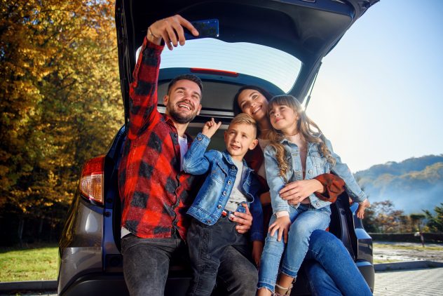 Happy parents with their children making selfie on smart phone while sitting in the trunk.